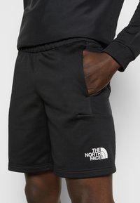 Personne portant un short de sport noir avec une taille élastique et le logo The North Face sur la jambe gauche, main dans la poche.