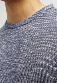 Texturerad marinblå tröja med rund halsringning. Materialet har ett stickat mönster med ljusa och mörka trådvariationer som skapar en fläckig effekt.