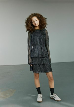 Fekete, réteges ruha hosszú, áttetsző ujjal és textúrált mintával. Fehér sportcipővel és fekete zoknival viselve.