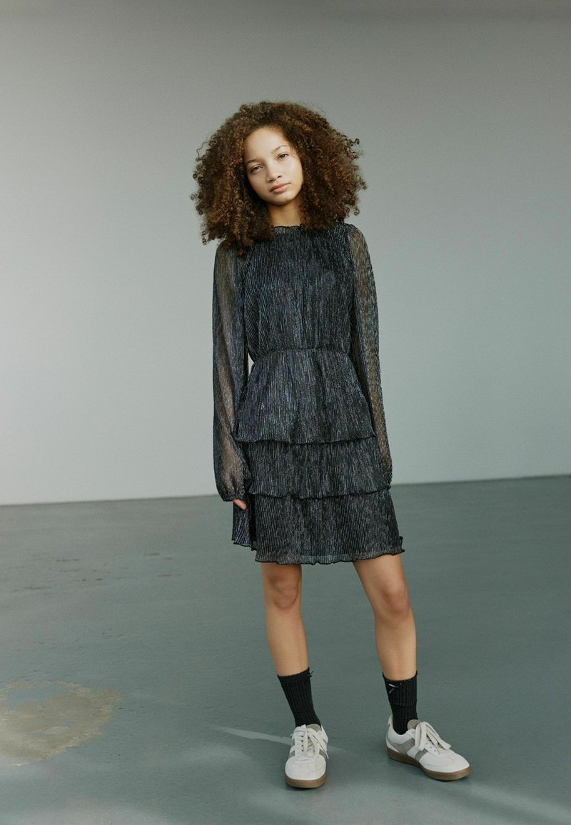Čierne šaty s viacerými vrstvami a dlhými priesvitnými rukávmi s textúrovaným vzorom. Nosené s bielymi teniskami a čiernymi ponožkami.