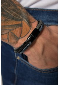Bracciale intrecciato in pelle nera e grigia con chiusura in metallo nero lucido, indossato su un polso decorato con un tatuaggio dettagliato, posizionato su un tessuto di denim.
