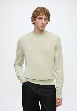 Junger Mann mit lockigem blondem Haar, der einen hellbeigen Langarmpullover und dunkle Hose trägt, mit einer Hand in der Tasche, vor weißem Hintergrund.