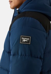 Giacca imbottita blu con inserti neri sulle spalle, colletto alto e patch con il logo Reebok sulla manica. Tessuto liscio e isolante.