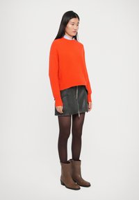 Maglione arancione a coste, gonna nera con zip, stivaletti marroni alla caviglia e collant scuri. Modella in piedi contro uno sfondo chiaro e semplice.