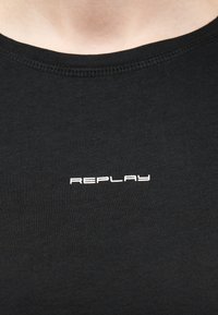 Zbliżenie czarnej koszulki z okrągłym dekoltem, z białym logo "REPLAY" umieszczonym na środku klatki piersiowej.