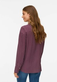 Kvinna med långt vågigt brunt hår som bär en vinröd och vit randig långärmad tröja och blå jeans, vänd åt sidan mot en vit bakgrund.
