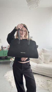 Naine mustas riietuses hoiab suurt musta käekotti heledas elutoas, kus on valge diivan, padjad, roheline kapp ja üleval lühter.