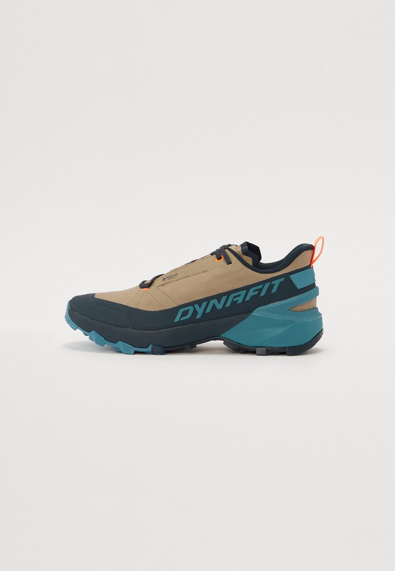 Trailrunning-Schuhe mit einem beigen Obermaterial und dunkelblauen Akzenten, die ein strukturiertes Design, eine Gummisohle und eine Zuglasche an der Ferse aufweisen.