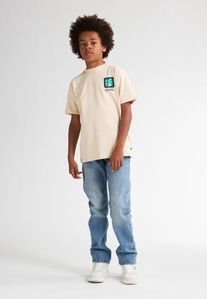 Camiseta de algodón beige con un logotipo cuadrado verde, de manga corta y un pequeño detalle de costura roja, combinada con jeans de mezclilla azul claro y zapatillas blancas.