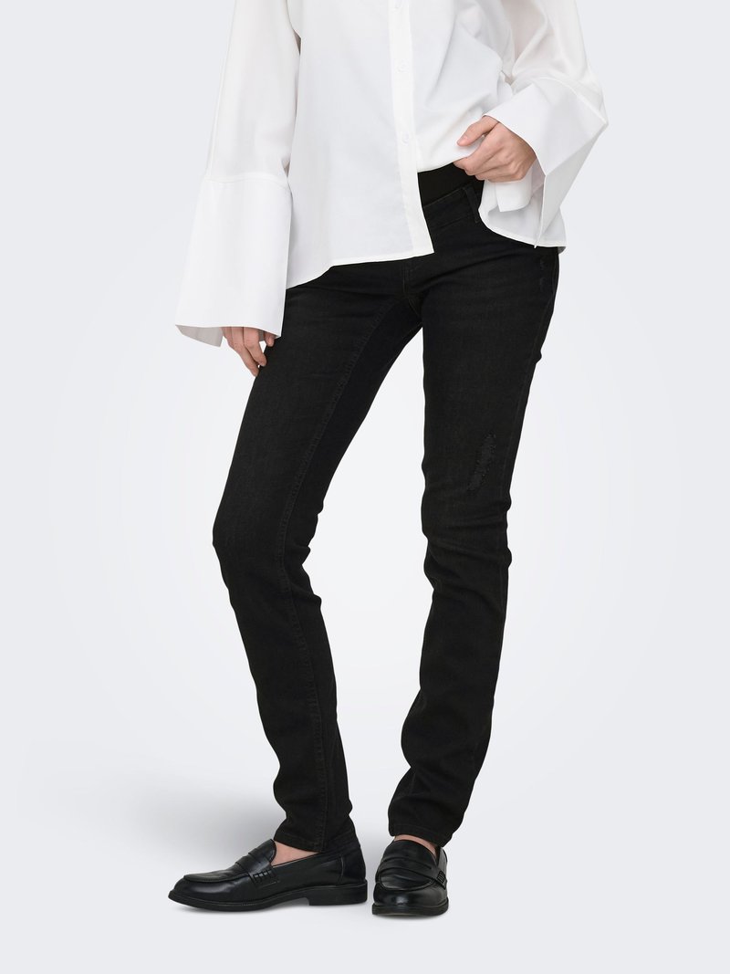 Personne portant un jean noir ajusté, des mocassins noirs et une chemise blanche à manches longues avec des poignets évasés, debout devant un fond uni.