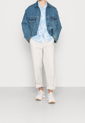 Homme portant une chemise bleue à motifs floraux, une veste en jean oversize, un pantalon cargo beige retroussé aux chevilles et des baskets blanches, debout les mains dans les poches.