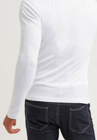 Camiseta blanca de manga larga con un diseño ajustado, textura suave y cuello redondo, combinada con jeans oscuros que tienen bolsillos traseros y detalles de costura.
