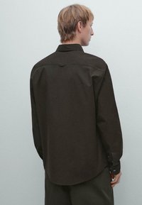 Persona con capelli biondi corti che indossa una camicia lunga e larga di colore marrone scuro e pantaloni scuri, girata di spalle contro uno sfondo chiaro e semplice.