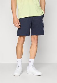 Marineblå trening shorts i myk stoff med elastisk midje, med hvit logo detalj langs siden, parret med hvite joggesko og sokker.
