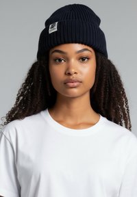 Laivastonsininen ribbineulottu pipo, jossa on tasainen päälliosa ja valkoinen merkitty merkki, jota käyttää henkilö yksivärisessä valkoisessa t-paidassa neutraalia taustaa vasten.