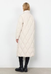 Lange, lichtbeige gewatteerde jas met hoge kraag, ruitpatroon en zijritsen. Draagt over zwarte leggings en chunky zwarte laarzen.