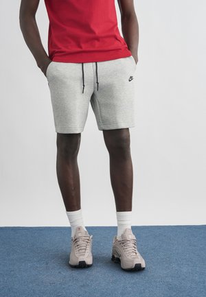 Asmuo, dėvintis pilką Nike šortus, baltas klasikinės stiliaus kojines, smėlio spalvos sportinius batelius ir raudoną marškinėlį, stovintis ant mėlyno kilimo priešais paprastą foną.