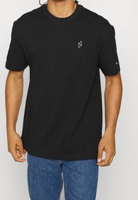 Czarna koszulka z okrągłym dekoltem, wykonana z bawełny, z krótkim rękawem. Posiada małe białe logo na piersi oraz luźny krój. Założona na niebieskie dżinsy.