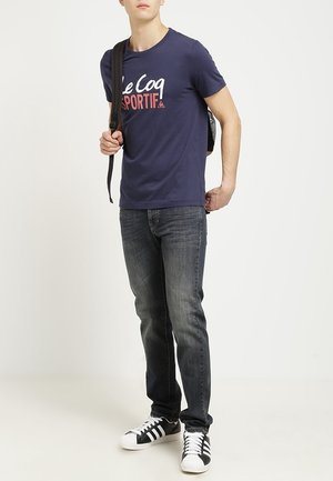 Navy blue t-shirt with white and red "Le Coq Sportif" text, paired with dark denim jeans and black-and-white sneakers. Black backpack worn.