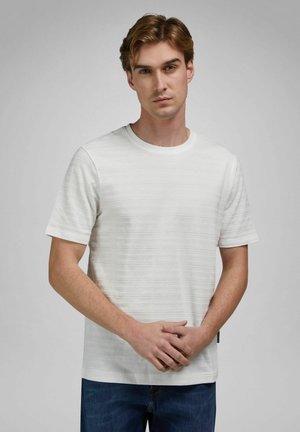 Giovane uomo con capelli castano chiaro che indossa una camicia bianca a maniche corte con trama e jeans blu, in piedi con le mani saldamente intrecciate.