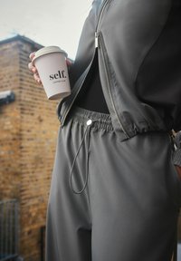 Gray athletic jacket with a zip closure, paired with matching gray joggers featuring an elastic waistband. Holding a cup labeled "self. coffee club."