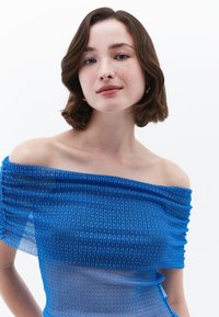 Modrá halenka s odkrytými rameny vyrobená z texturovaného materiálu s vertikálními pruhy a nabíraným designem na rukávech a hrudníku. Viditelné stříbrné detaily.