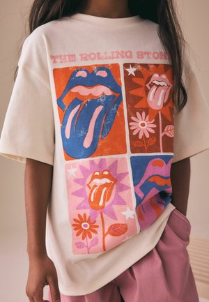 Dieťa nosiace biele tričko Rolling Stones s farebnými motívmi jazyka a pier, ružové šortky, ruka v vrecku.