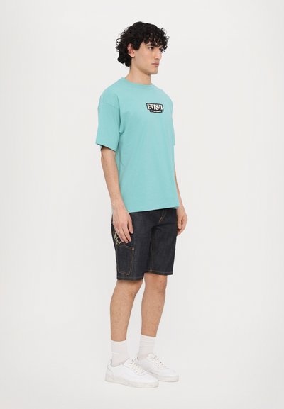 Τυρκουάζ βαμβακερή μπλούζα με μαύρο λογότυπο, συνδυασμένη με σκούρα τζιν σορτς που διαθέτουν πλαϊνές τσέπες και ανοιχτό ραφές; λευκά αθλητικά παπούτσια.