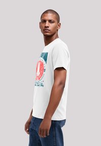White cotton t-shirt featuring a colorful graphic design with text, palm trees, and a circular emblem, paired with blue denim jeans.