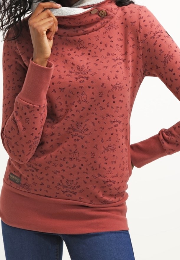 Femme portant un sweat-shirt couleur rouille avec des motifs d'animaux et de plantes de la forêt, avec un col haut boutonné et des manches longues, associé à un jean.