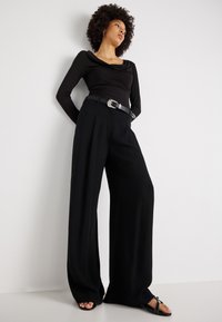 Črn dolg rokav z drapiranim dekoltejem, v kombinaciji s širokima črnima hlačami in črnim pasom s srebrnimi detajli. Črne sandale dopolnjujejo videz.