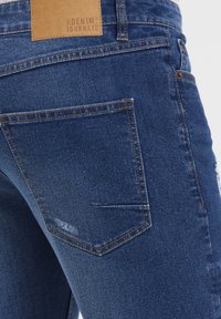 Blå denimjeans med falmet tekstur, med en brun lærlapp og kontrasterende søm, som viser to baklommer.