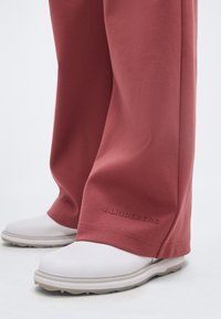 Άσπρα παπούτσια συνδυασμένα με κόκκινο παντελόνι που έχει το λογότυπο J.Lindeberg κοντά στα τελειώματα, στέκονται σε φωτεινή επιφάνεια.