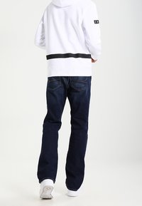 Bílý sweatshirt s kapucí a černým vodorovným pruhem v pase, v kombinaci s tmavě modrými džíny a bílými teniskami.