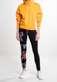 Oranžová mikina s kapucí a stahovacími šňůrkami, zkombinovaná s černými legínami s barevnými poti štěnými grafikami a bílými teniskami.