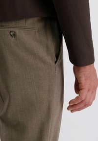 De hand van een man naast de olijfgroene broekzak van een pantalon met een knoopzak aan de achterkant, terwijl hij een donkerbruin, gestructureerd blazerjack draagt.