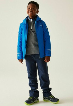Ragazzo sorridente che indossa una giacca impermeabile blu, un maglione grigio con zip, pantaloni blu navy e scarpe da ginnastica nere e verdi, in piedi davanti a uno sfondo chiaro.