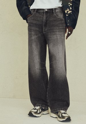 Personne portant un jean noir délavé à jambes larges, des baskets blanches avec des accents noirs et beiges, une chemise blanche et une veste noire avec un motif camouflage.