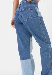 Mujer vestida con unos vaqueros de cintura alta de patchwork en tonos de azul con paneles de denim claro y oscuro, con la mano descansando cerca de la cadera, contra un fondo blanco.