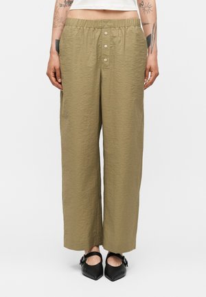 Χακί παντελόνια με φαρδιά μπατζάκια, ελαστική μέση και κουμπωτό πέτο, φορεμένα με μαύρα μυτερά παπούτσια τύπου Mary Jane.