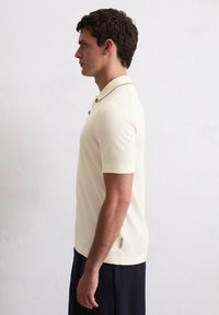 Kremowa koszulka polo z krótkim rękawem o teksturowanym wykończeniu, z dwoma guzikami i bocznym rozcięciem na dolnym brzegu.