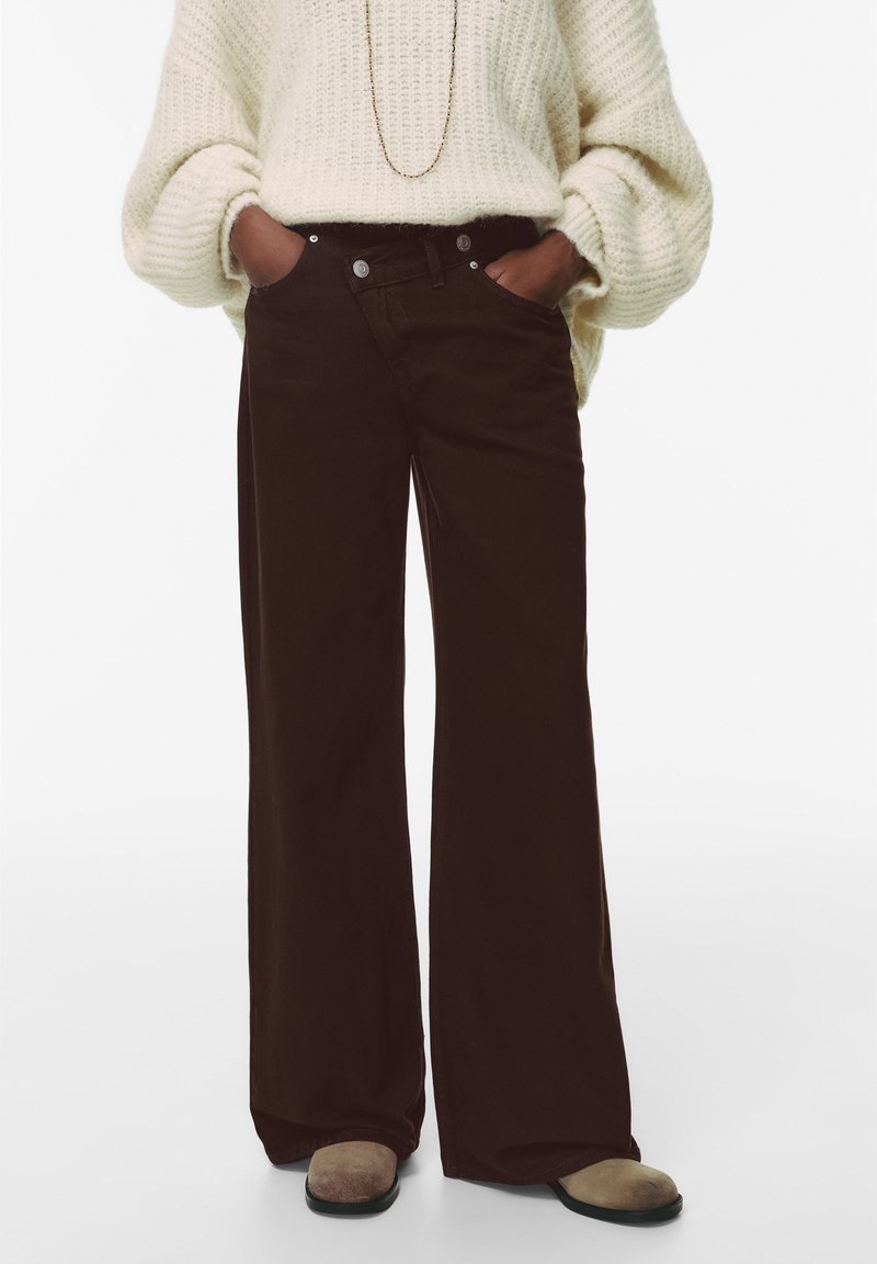 Pantaloni a gamba larga in corduroy marrone con vita alta, caratterizzati da un design a cinque tasche e bottoni metallici a contrasto, abbinati a un maglione in maglia a trecce.