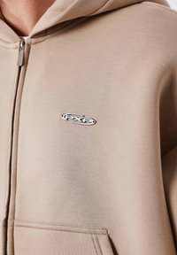 Felpa beige con zip realizzata in tessuto morbido, con logo argentato sul petto, tasca a marsupio e cappuccio con coulisse.