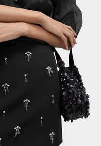 Schwarze Pailletten-Handtasche mit runder Form, metallischen Hardware und strukturierten Verzierungen. Schwarzer Rock mit Kristalldetails und Verzierungen.