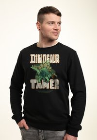 Felpa nera con una grafica che presenta un dinosauro verde e la scritta "Domatore di Dinosauri". Polsini e orlo a coste, texture morbida.