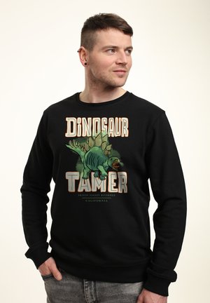 Zwarte sweatshirt met een graphic van een groene dinosaurus en de tekst "Dinosaur Tamer." Ribgebreide manchetten en onderrand, zachte textuur.
