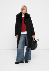 Cappotto nero con colletto in pelliccia sintetica, maglione rosso a maglia, jeans a gamba larga in denim e una borsa nera. Modella in piedi contro uno sfondo neutro.