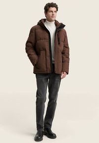 Rjava puhovka z kapuco, z zadrgo spredaj, bočnimi žepi in črnimi poudarki. Oblečena čez belo oprijeto majico z visokim ovratnikom in sive kavbojke.