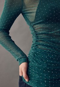 Langærmet bluse i grønblå med tekstureret, gennemsigtigt stof med lodrette striber og spredte rhinsten; har rynkede detaljer ved taljen.