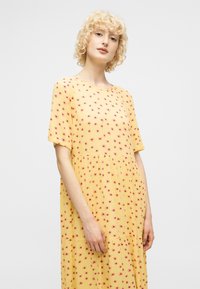 Ξανθός άνθρωπος με σγουρά μαλλιά που φοράει μια άνετη κίτρινη φόρεμα με κοντά μανίκια καλυμμένη με μικρά ροζ λουλουδάτα σχέδια, στέκεται μπροστά σε ένα απλό φόντο.